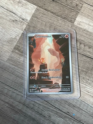 Pokémonkort Charmander & Venusaur GX - Två Pokémonkort: ett med Charmander i tecknad stil med bruna och orange toner, samt ett holografiskt Venusaur & Snivy GX TAG TEAM-kort med färgglad glans. Båda korten är i plastfickor och har detaljerade illustrationer och text på engelska. Perfekt för samlare eller Pokémonfans.
