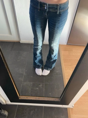 Blå bootcut jeans med låg midja 25/32 - Snygga blå jeans med bootcut och låg midja, klassisk femficksmodell och dubbla silverknappar fram. Jeansen har kontrastsömmar och är tillverkade i slitstarkt denim. Perfekt passform för dig som gillar retrovibbar och markerad siluett. De är stretchiga och passar större och mindre storlekar