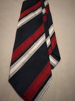 Randig slips i rött, vitt och marinblått - Snygg slips med diagonala ränder i marinblått, rött och vitt. Tillverkad i ett glansigt tyg som känns som siden. Perfekt accessoar för att lyfta din outfit med en klassisk och stilren look.