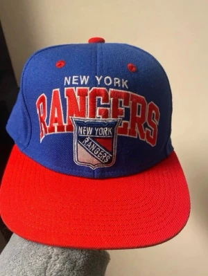 New York Rangers  - Snygg blå och röd snapback keps från Mitchell & Ness med New York Rangers broderat framtill och logga. Justerbar baktill med röd plastrem. Klassisk sportig look med platt skärm och broderade detaljer. Perfekt för dig som gillar hockey och streetwear.