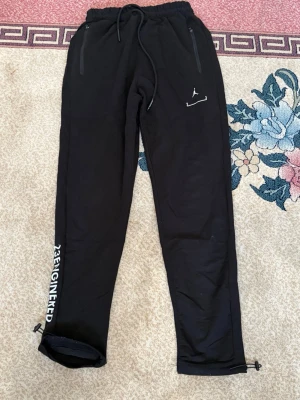 Svarta Jordan mjukisbyxor 23 Engineered - Säljer ett par svarta mjukisbyxor från Jordan med vita detaljer och texten '23 ENGINEERED' längs benet. Byxorna har dragsko i midjan, dragkedjefickor och justerbara benslut. Perfekta för chill eller streetwear.