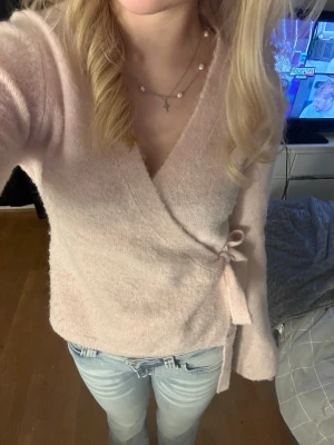 Ljusrosa omlottstickad tröja - Supersöt ljusrosa stickad omlott-tröja med knytband i sidan. Tröjan har en djup v-ringning och långa ärmar. Perfekt för dig som gillar en mjuk och feminin stil. Materialet ser ut att vara mjukt och fluffigt, troligen en blandning med akryl eller liknande.(köp med Instabox‼️)