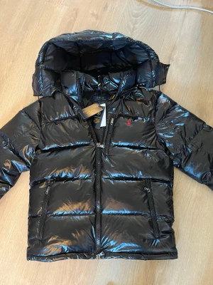 Svart pufferjacka från Polo Ralph Lauren - Snygg svart pufferjacka från Polo Ralph Lauren med glansig finish och huva. Jackan har två dragkedjeförsedda fickor, hög krage och ikoniska röda loggan broderad på bröstet. Perfekt för kalla vinterdagar och riktigt trendig look.
