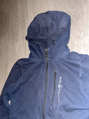 Marinblå hoodie från Sail Racing - Snygg marinblå hoodie från Sail Racing med dragkedja framtill och en extra bröstficka med dragkedja. Hoodien har justerbar huva och logga tryckt på bröstet. Tillverkad i mjukt material som känns skönt mot huden. Perfekt för en avslappnad och sportig stil.