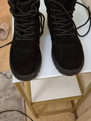 Svarta mockakängor med snörning - Säljer ett par svarta kängor i mocka med grov sula och snörning framtill. De har en vadderad kant upptill för extra komfort och en klassisk, stilren look som passar till många outfits. Perfekta för dig som gillar en tuffare stil.