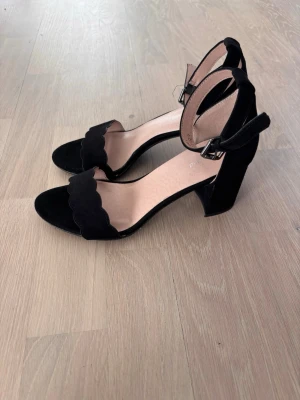 Svarta mocka-pumps med blockklack - Stilrena svarta pumps med öppen tå, bred blockklack och justerbart ankelband. Främre remmen har en snygg vågig kant och skon är i mockaimitation. Perfekt för dig som vill ha en klassisk men trendig look. Köpta på Deichmann men från märket Graceland. Använda en gång men har små slitningar som man ser på bilderna. På sista bilden ser man att innersulan sitter lite löst men skulle gissa på att de enkelt går att limma ihop. Hör av dig om du har några frågor💕