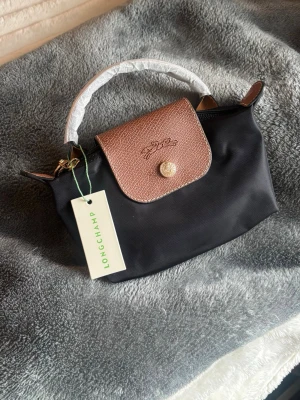 Svart handväska Mini Longchamp - Snygg svart handväska från Longchamp med brunt lock i präglat läder och guldfärgade detaljer. Väskan har ett kort handtag och dragkedja upptill. Axelrem tillkommer❤️ Helt ny! 