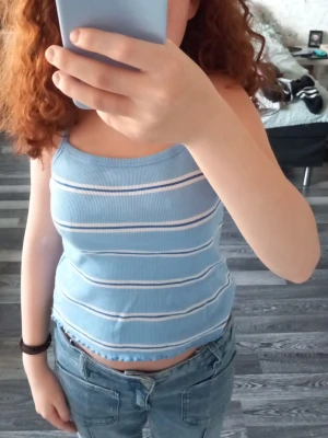 Blåvit randig ribbad linnetopp - Säljer en ljusblå ribbad linnetopp med vita och ljusrosa ränder. Toppen har smala axelband och en croppad passform med vågig kant nertill. Perfekt för sommaren och enkel att matcha med jeans eller kjol.