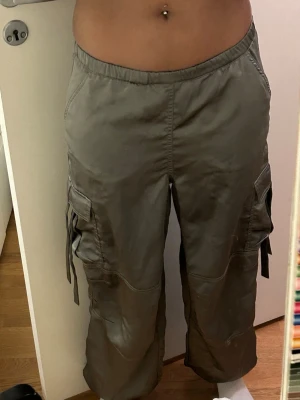 Grön/gråa cargobyxor med fickor - Säljer ett par grön/grå cargobyxor med bred resår i midjan och flera stora fickor på sidorna och baktill. Byxorna har avslappnad passform och snördetaljer på benen. Perfekta för en streetwear-look och riktigt sköna att bära.