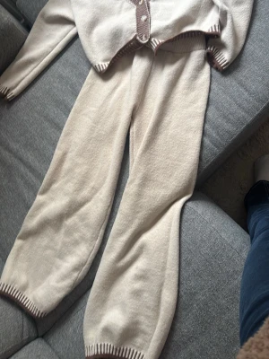 Beige loungewear-set med bruna detaljer - Mysigt stickat loungewear-set i beige med bruna ribbade detaljer. Setet består av en långärmad kofta med knappar och matchande byxor med resår i midja och benslut. Perfekt för chill dagar hemma. Materialet är mjukt och stickat för extra komfort.