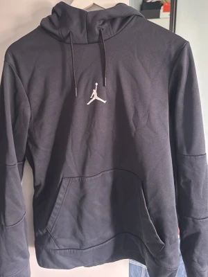 Svart Jordan hoodie med logga - Snygg svart hoodie från Jordan med den klassiska Jumpman-loggan broderad i vitt på bröstet. Hoodien har huva med dragsko och en stor känguruficka framtill. Tillverkad i mjukt bomullsmaterial som är perfekt för chill eller streetwear.