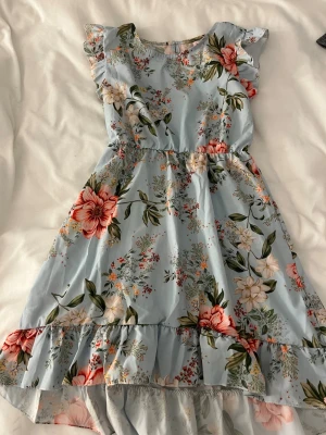 Blommig ljusblå volangklänning - Söt ljusblå klänning med stora blommor i rosa, vitt och grönt. Klänningen har volangdetaljer vid ärmar och nederkant, rund halsringning och kort ärm. Perfekt för sommarens alla tillfällen och ger en romantisk vibe.