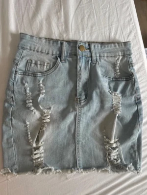 Ljusblå jeanskjol med slitningar - Trendig ljusblå jeanskjol med slitna detaljer framtill och rå kant nertill. Kjolen har klassiska fem fickor, bälteshällor och knappstängning. Perfekt för dig som gillar en avslappnad och cool stil.