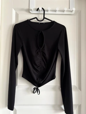 Svart långärmad topp med knyt - Snygg svart långärmad topp med rund halsringning och en unik cut-out framtill. Toppen har rynkade detaljer längs mitten och ett knyt i nederkanten som ger en cool, croppad look. Perfekt att styla med höga jeans eller kjol.