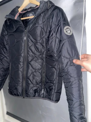 Svart pufferjacka från Napapijri - Snygg svart pufferjacka från Napapijri med quiltat mönster och huva. Jackan har dragkedja framtill, två sidofickor med dragkedja och en rund logotyp-patch på ärmen. Perfekt för kalla dagar och riktigt skön att bära.
