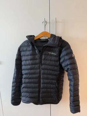 Svart dunjacka från Peak Performance - Snygg svart dunjacka från Peak Performance med huva och dragkedja framtill. Jackan har en quiltad design med horisontella sömmar och logga på bröstet samt ärmen. Perfekt för kalla vinterdagar och riktigt lätt att bära.