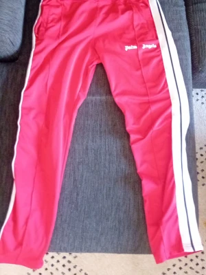 Palm Angels  - Säljer ett par röda mjukisbyxor från Palm Angels med vita sidoränder och svart detalj. Byxorna har en avslappnad passform och är tillverkade i ett glansigt, sportigt material. Loggan är tryckt i vitt på vänster sida. Perfekta för en streetwear-look.