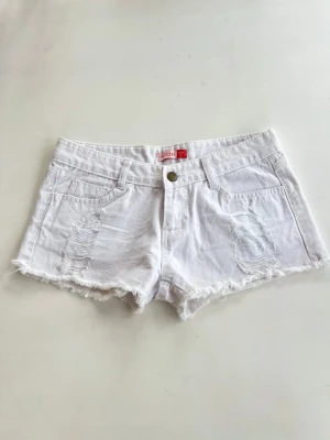 Vita jeansshorts med låg midja  - Säljer ett par vita jeansshorts från blue ray med slitna detaljer framtill och på bakfickorna. Shortsen har råa kanter nertill, klassisk femficksmodell och knappgylf. Perfekta för sommaren och ger en avslappnad vibe.