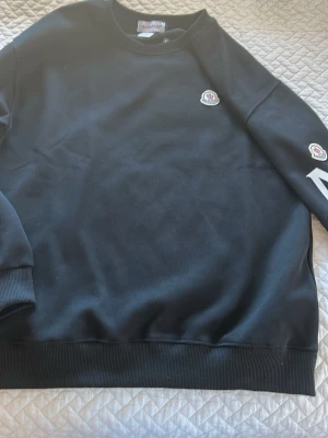 Svart sweatshirt från Moncler - Säljer en svart sweatshirt från Moncler med rund halsringning och ribbade muddar. Tröjan har Moncler-logga på bröstet och ärmen, samt en enkel och clean design. Perfekt för dig som gillar stilrena plagg med exklusiv känsla.