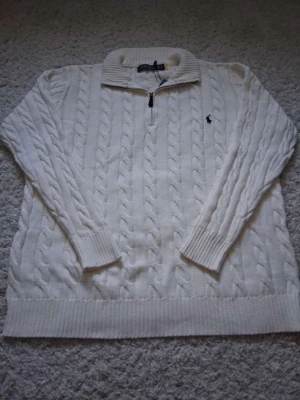 Vit kabelstickad tröja Polo Ralph Lauren - Vit kabelstickad tröja från Polo Ralph Lauren med halv dragkedja och hög krage. Tröjan har ribbade muddar och nederkant samt den klassiska broderade loggan på bröstet. Perfekt för lager-på-lager och snygg till jeans.