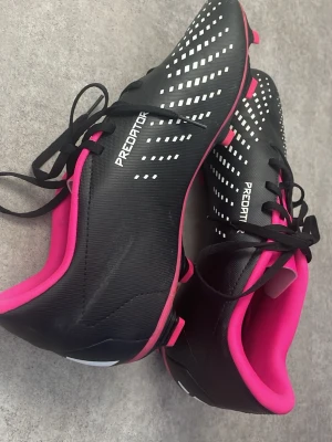 Adidas Predator fotbollsskor - Snygga Adidas Predator fotbollsskor i svart med vita detaljer och rosa insida samt dobbar. Skorna har klassisk snörning och är tillverkade i syntetmaterial för bra grepp och kontroll på planen. Perfekta för dig som vill sticka ut med stil på matchen.