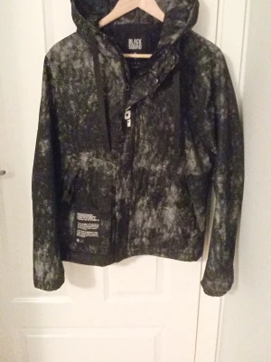 Jacka - Cool vindjacka från Black Squad i svart och vitt mönster med tryck på ryggen. Jackan har huva, dragkedja och snörning samt tryckdetaljer framtill. Perfekt för dig som vill sticka ut med streetstyle och sportig look.