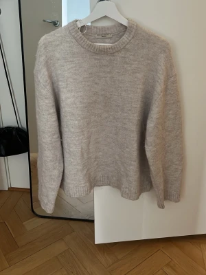 Beige stickad tröja Gina Tricot - Mysig beige stickad tröja från Gina Tricot med rund halsringning och ribbade muddar vid ärmslut och nederkant. Tröjan har en avslappnad passform och är tillverkad i ett mjukt material som passar perfekt till kyligare dagar.