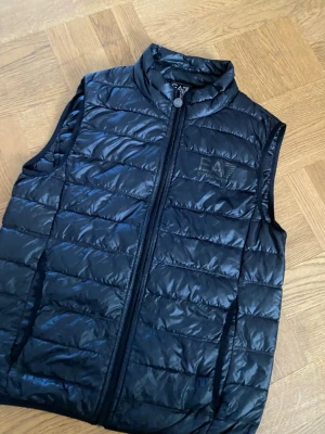Svart dunväst från Emporio Armani - Snygg svart dunväst från Emporio Armani EA7 med quiltad design och hög krage. Västen har dragkedja framtill och diskret logga på bröstet. Perfekt för lager på lager och ger en sportig vibe till din outfit. Lite hål som har jag har fixat su kan se de på bilden men man ser den inte.