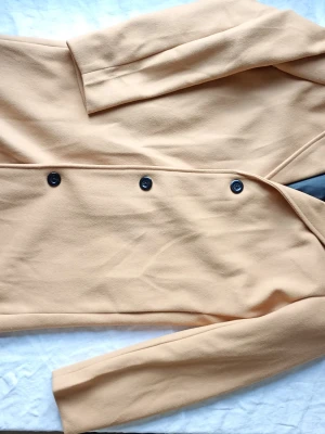 Beige dubbelknäppt kappa - Stilren beige kappa med dubbelknäppning och svarta knappar. Klassisk rak passform och långa ärmar. Perfekt för dig som gillar minimalistisk och tidlös stil. Materialet ser ut att vara mjukt och något tjockare, vilket ger en snygg siluett.