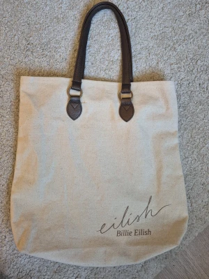 Billie eilish väska - Stilren shoppingväska i beige canvas med mörkbruna handtag i konstläder. Tryck med 'eilish Billie Eilish' på framsidan. Perfekt för dig som vill ha en rymlig och trendig väska med artistkänsla.