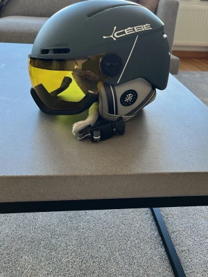 Cébé hjälm med gult visir - Cool hjälm från Cébé i matt mörkgrå färg med integrerat gult visir. Hjälmen har mjuka, grå öronlappar och justerbart hakband. Perfekt för dig som vill ha både skydd och stil på vintern. Materialet är hårdplast med vadderade insidor för extra komfort. Storlek M