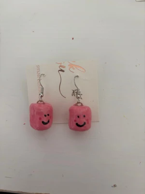 Rosa örhängen med smiley - Unika handgjorda örhängen med rosa pärlor formade som små marshmallows med glada smileys. Hängena har metallkrokar och en lekfull design som sticker ut. Perfekta för dig som gillar färgglada accessoarer och vill ha något annorlunda.
