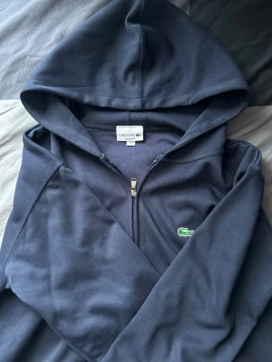 Mörkblå hoodie från Lacoste - Snygg mörkblå hoodie från Lacoste med dragkedja framtill och klassisk krokodil-logga på bröstet. Hoodien har huva och två fickor framtill. Tillverkad i mjukt material som känns skönt mot huden. Perfekt för en avslappnad och stilren look.