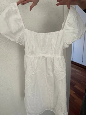 Vit kort klänning  - Söt vit kort klänning från H&M Divided med puffärmar och smockdetaljer upptill. Klänningen har en luftig passform och är tillverkad i mjuk bomull, perfekt för varma dagar. Enkel och stilren design som är lätt att matcha med accessoarer. Aldrig använd, storlek M💕