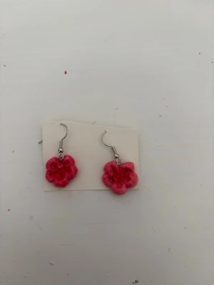 Rosa blommiga örhängen - Säljer ett par handgjorda örhängen i form av rosa blommor. De har krokar i silverfärg och själva blomman är i en glansig, ceriserosa nyans med detaljerade blomblad. Perfekta för dig som vill ha något färgglatt och unikt till din stil.