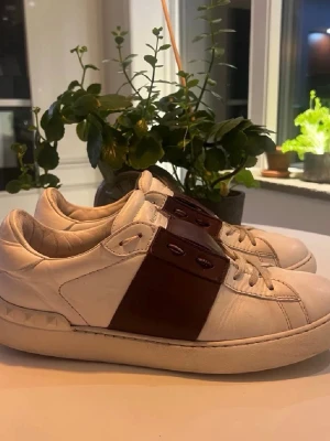 Valentino vita sneakers med brun detalj - Snygga vita sneakers från Valentino med bred brun läderdetalj över snörningen och nitar på hälen. Klassisk låg modell med rund tå och vita skosnören. Perfekta för dig som vill ha en stilren men unik look.