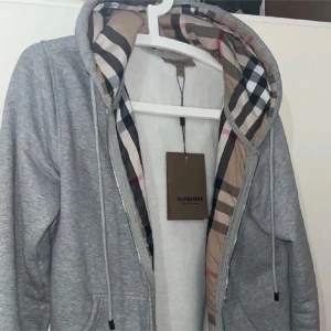 Grå hoodie från Burberry med huva - Snygg grå zip-hoodie från Burberry med klassiskt rutigt foder i huvan. Hoodien har dragsko i huvan, fickor framtill och dragkedja. Perfekt för dig som vill ha en stilren och exklusiv look med ikoniska Burberry-detaljer.