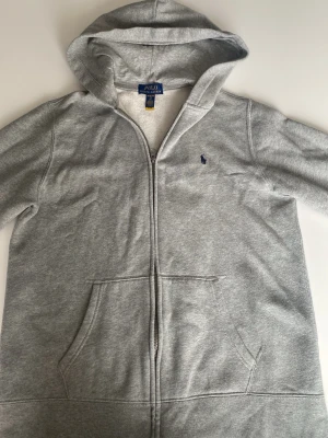 Grå hoodie från Polo Ralph Lauren - Klassisk grå hoodie med dragkedja från Polo Ralph Lauren. Mjuk bomullsblandning, två fickor framtill och broderad marinblå logga på bröstet. Inte använd på grund av att den var för liten. 