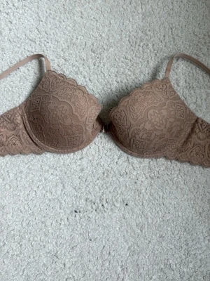 Brun spets-bh med vaddering 75b - Snygg brun bh med vadderade kupor och vacker spetsdetalj över hela bh:n. Kuporna har scallop-kant och tunna axelband. Perfekt för dig som gillar en feminin och stilren look. Ingen synlig logga eller märke.