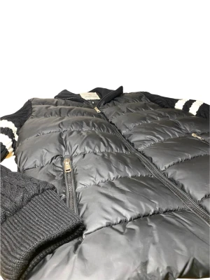 Svart dunväst från Moncler - Snygg svart dunväst från Moncler med stickade ärmar och vita ränder på ärmarna. Västen har dragkedja framtill, två fickor med dragkedja och ribbade muddar. Perfekt för lager-på-lager och ger en sportig vibe.