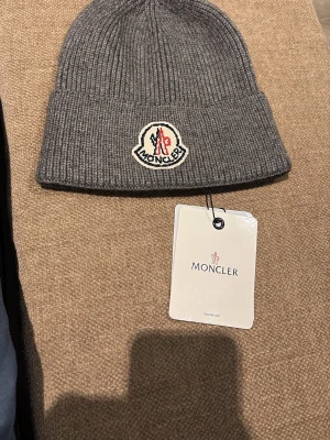 Grå ribbstickad mössa från Moncler - Snygg grå ribbstickad mössa från Moncler med uppvikt kant och Moncler-logga framtill. Tillverkad i mjukt material som känns lyxigt och passar perfekt för kalla dagar. Klassisk och stilren design som funkar till det mesta. Helt ny aldrig använd, skickas samma dag eller dagen efter köpet 