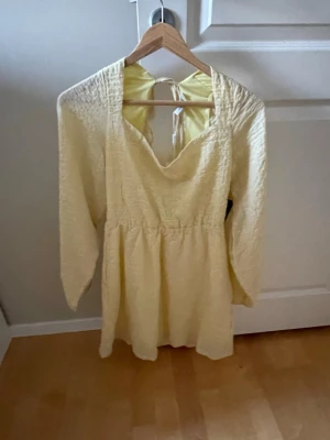 Ljusgul kort klänning med öppenrygg  - Söt ljusgul kortklänning med lång ärm och smockad överdel. Klänningen har en djup v-ringning och öppen rygg med knytband i nacken. Perfekt för varma dagar och ger en romantisk känsla.