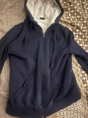 Mörkblå hoodie med fluffigt foder - Mysig mörkblå hoodie med dragkedja och stora fickor framtill. Insidan är fodrad med vitt, supermjukt teddyfoder som gör den extra varm. Huvan har dragsko och hela hoodien har en avslappnad passform. Perfekt för kalla dagar när du vill vara både bekväm och stilren.