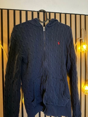 Mörkblå stickad hoodie från Polo Ralph Lauren - Mörkblå stickad hoodie med dragkedja från Polo Ralph Lauren. Tröjan har kabelstickat mönster, fickor framtill och en liten röd logga broderad på bröstet. Perfekt för dig som gillar klassisk och avslappnad stil. Säljer den pågrund av den är för liten, köpt för 1700kr
