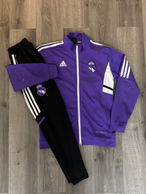 💜Lila Adidas Real Madrid träningsset(Barn)💜 - Säljer ett snyggt träningsset från Adidas x Real Madrid. Jackan är lila med vita och svarta detaljer, dragkedja och klubbmärke på bröstet. Byxorna är svarta med vita ränder och Real Madrid-logga. Perfekt för fotbollsträning eller chill. Passar till dig som är ca 150-160. 