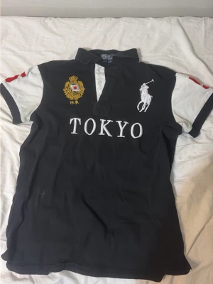 Svart piké Tokyo från Ralph Lauren - Snygg svart pikétröja från Ralph Lauren med vita ärmar och stora broderade detaljer. Framsidan har Tokyo-text, logga och emblem, medan baksidan har en stor röd siffra 5. Klassisk krage och knappar framtill.