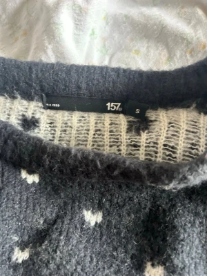 Mörkgrå stickad tröja från 157 - Mysig mörkgrå stickad tröja från 157 med vitt och rött nordiskt mönster över bröstet och små vita detaljer över hela tröjan. Tröjan har rund halsringning och långa ärmar. Perfekt för kalla dagar och ger en klassisk vinterlook.