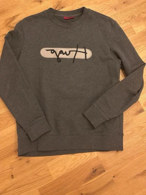 Mörkgrå sweatshirt från Hugo boss - Mörkgrå sweatshirt med rund halsringning och svart/vitt grafiskt tryck på bröstet. Tröjan har långa ärmar, ribbade muddar och är gjord i mjukt bomullsmaterial. Perfekt för en avslappnad och stilren look.