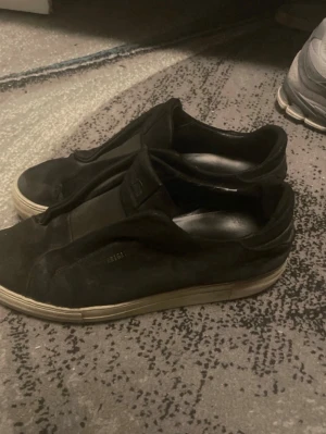 Svarta slip-on sneakers från Axel Arigato - Svarta slip-on sneakers från Axel Arigato i mocka med diskret logga på sidan. Skorna har en tjock vit sula och minimalistisk design utan snörning. Perfekta för en clean och stilren look.