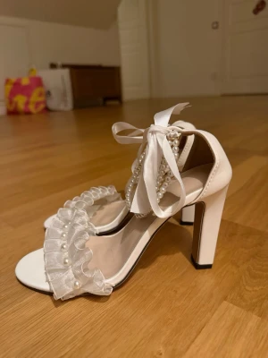 Vita pumps med pärlor och rosetter - Säljer ett par vita pumps med bred klack, dekorerade med pärlor och volang i organza framtill. Säljer då de ej kommer till användning. Endast använda 1 gång.